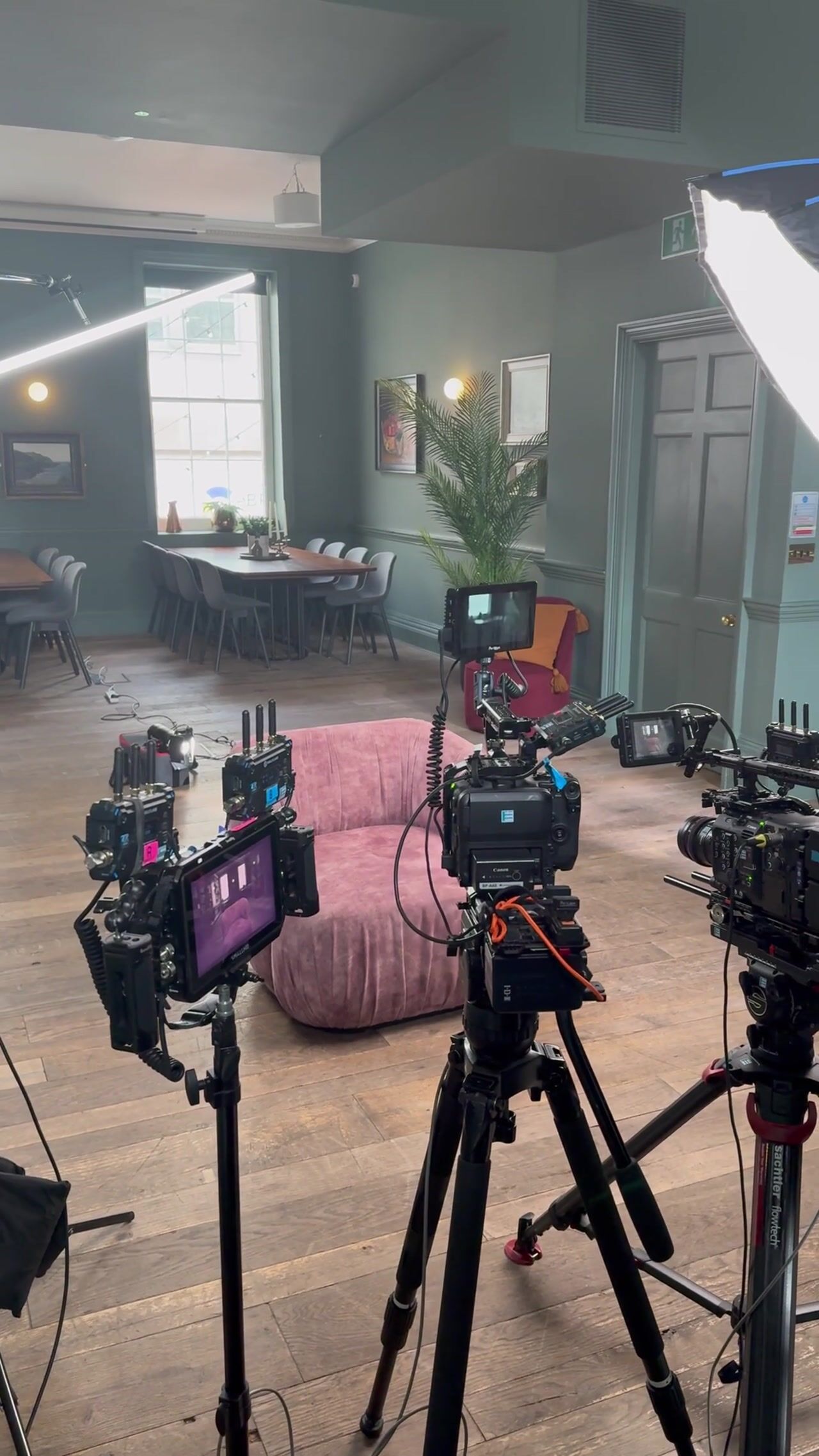 Camera monitors and tripod-mounted cameras face an empty pink armchair in a green-panelled room, with a softbox light and dining table in the background.