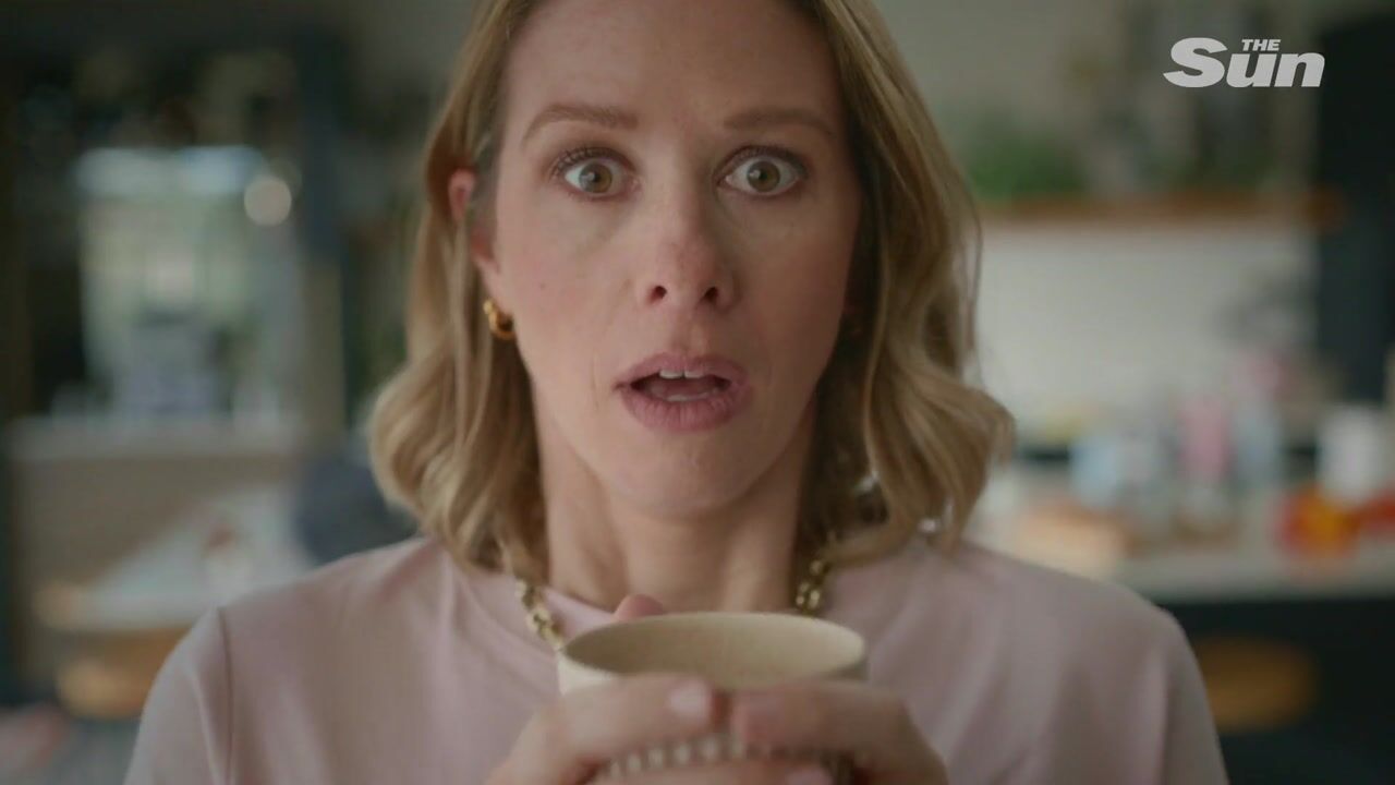 Close-up of a woman with wavy blonde hair, in a pink top, holding a mug, facing camera with a startled expression.