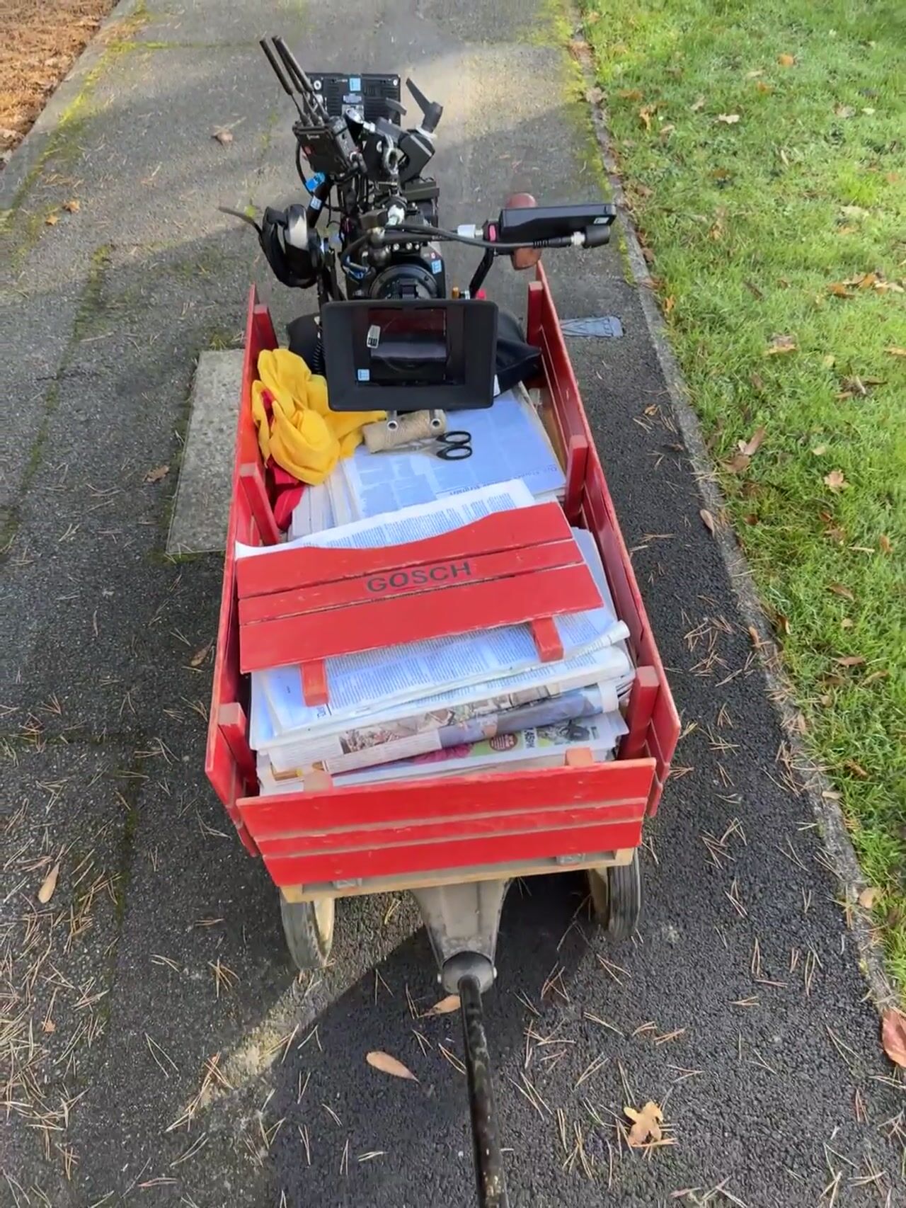 A red wagon marked "Gosch" loaded with folded newspapers, twine, a yellow cloth and a camera monitor rig, on a pavement beside grass.