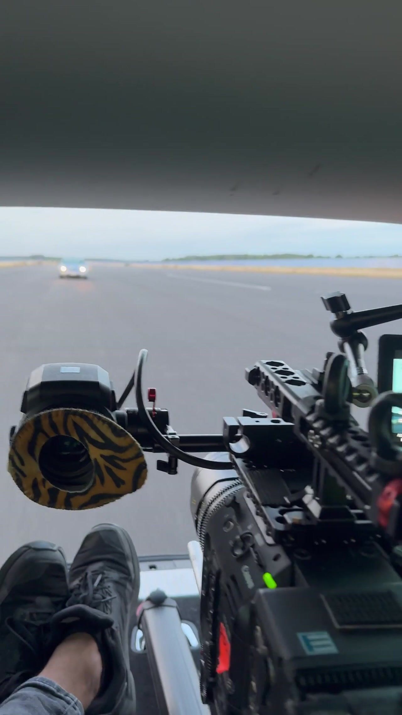 Camera rig with lens mounted on a car bonnet, filming another vehicle ahead on an open runway, crew member's feet in frame.