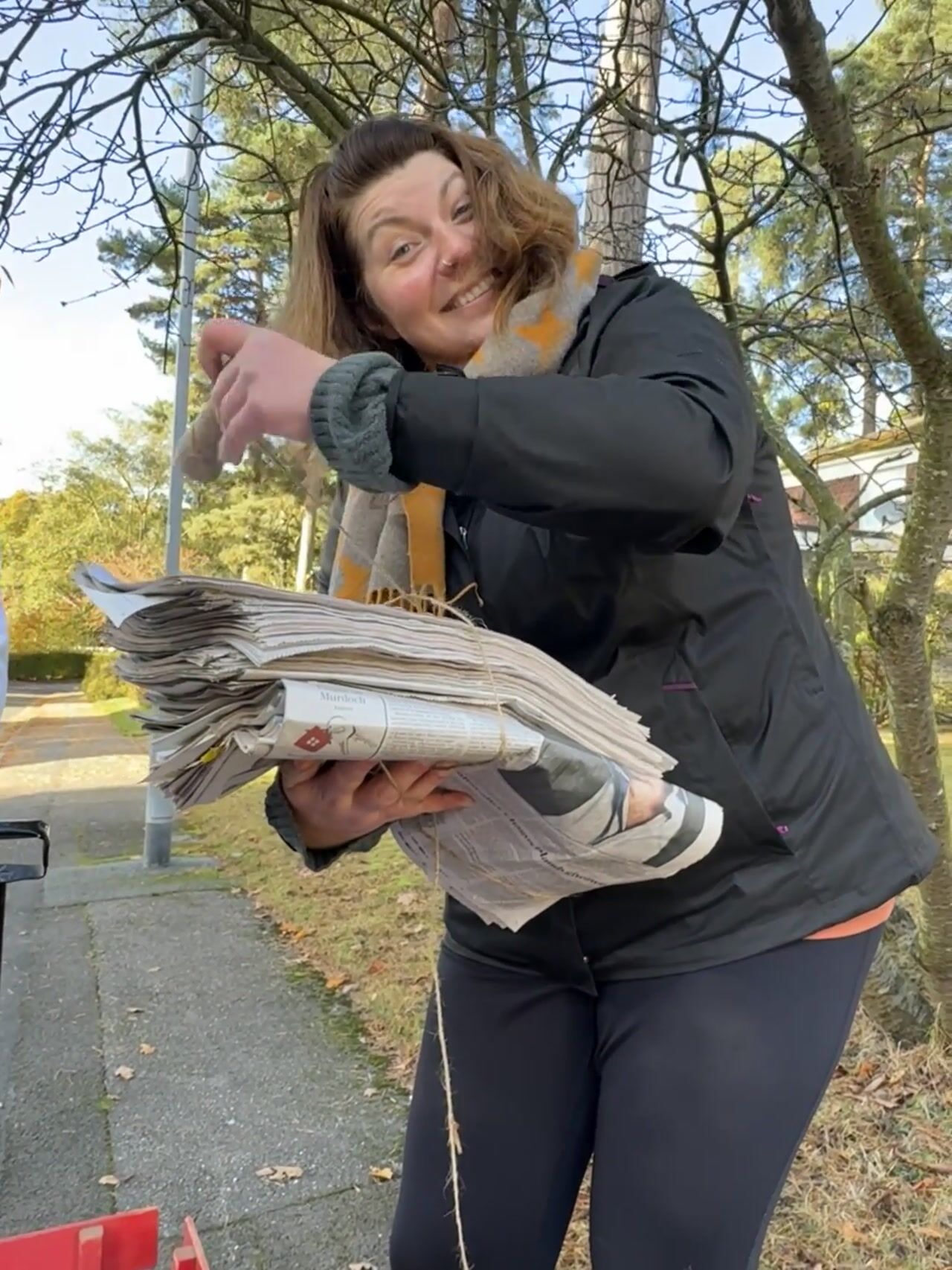 A woman with reddish-brown hair, in an orange-patterned scarf and dark jacket, smiles while holding a bundle of tied newspapers outdoors.
