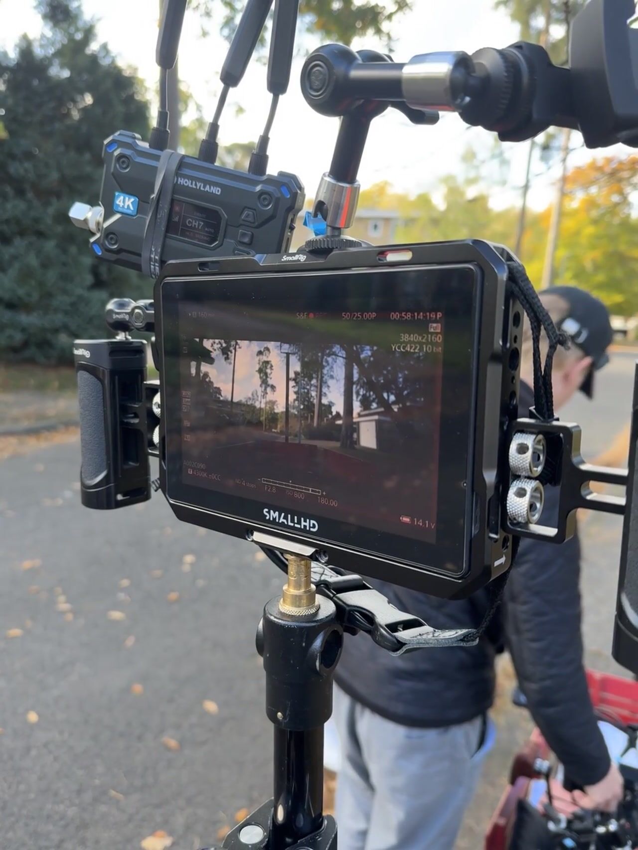 A camera monitor on a tripod displays a tree-lined street scene, with a wireless receiver mounted above, a man in a dark cap standing beside it outdoors.