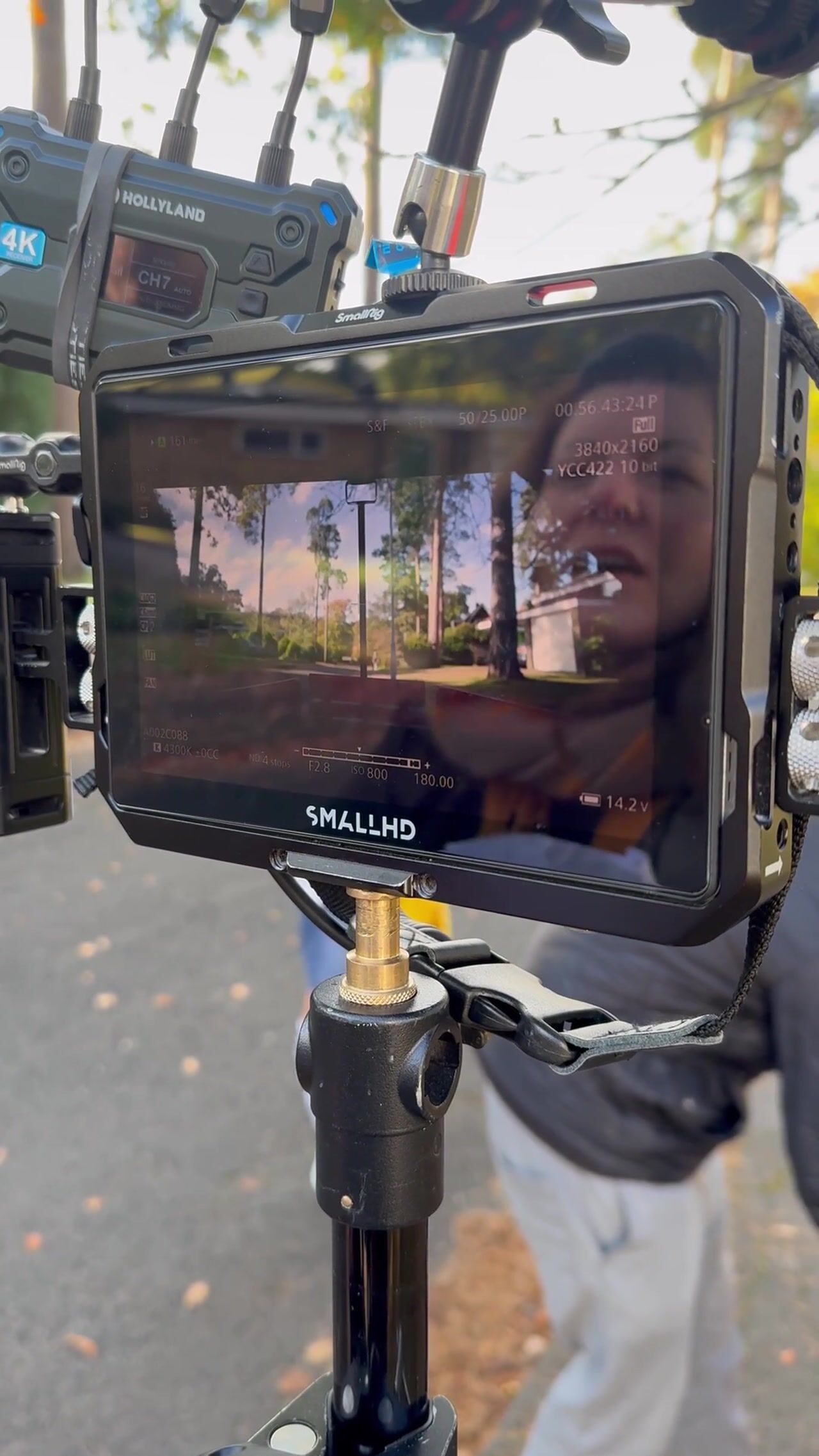A camera monitor on a tripod shows a tree-lined street, reflecting the blurred face of a dark-haired person behind it outdoors.