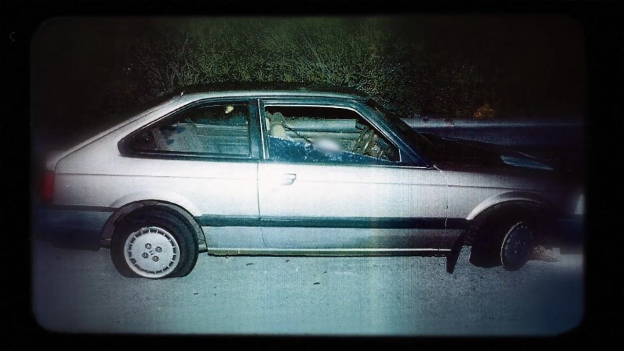 A pale saloon car parked side-on at night, its side window smashed and interior visible, with trees in the background.