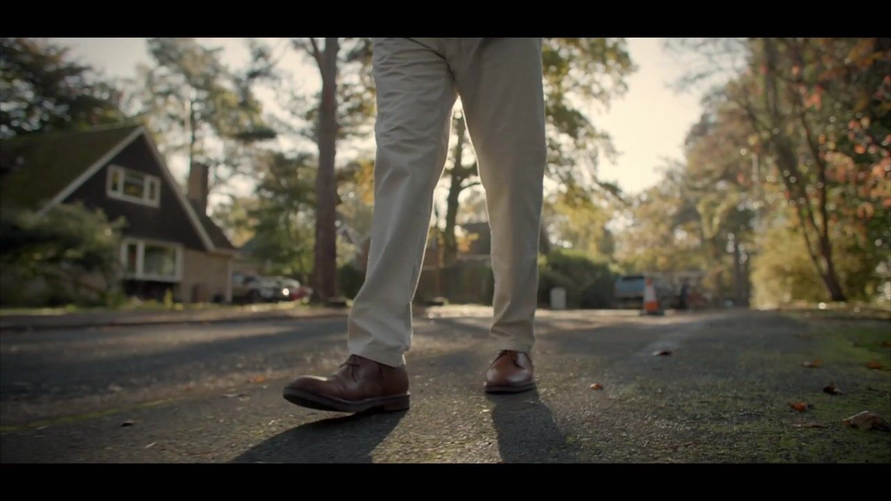 Low-angle close-up of a person's legs in beige trousers and brown shoes, walking along a suburban street lined with trees and houses.