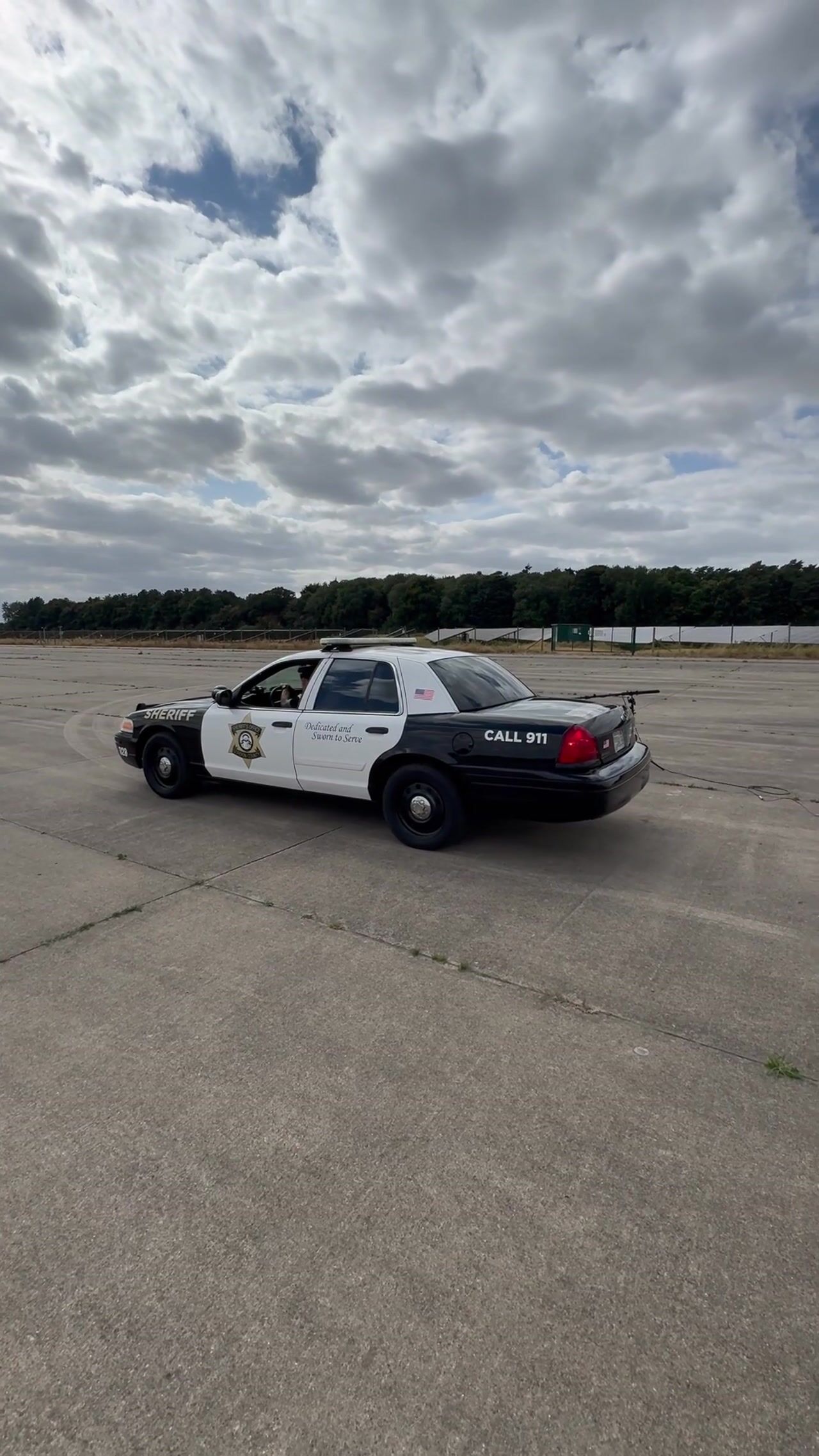 Black-and-white sheriff's saloon marked "Call 911" parked on an empty airfield, driven side-on beneath a wide cloudy sky.