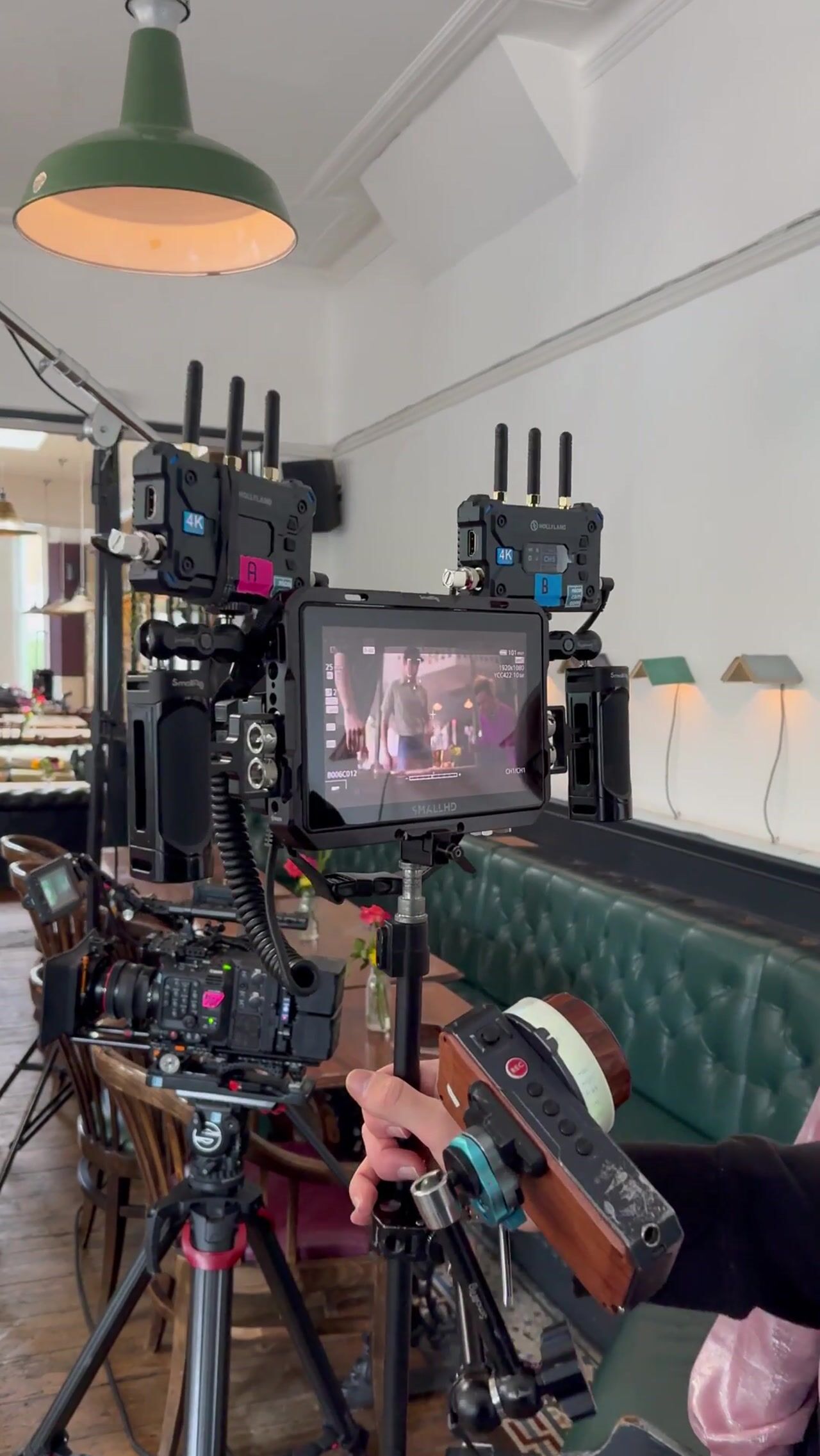 A cinema camera rig with monitor and wireless receivers on a tripod, its screen showing a recreated pub scene, a hand adjusting the focus wheel in the foreground.