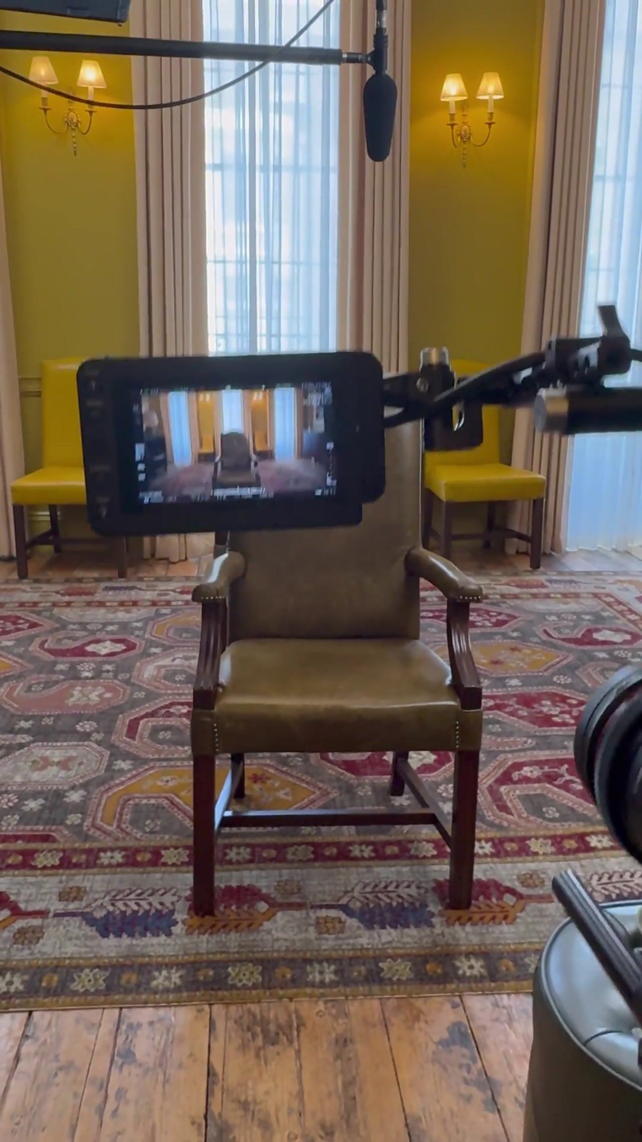 An empty leather armchair set up for an interview, with a camera monitor, boom microphone and yellow chairs in a grand room with yellow walls.