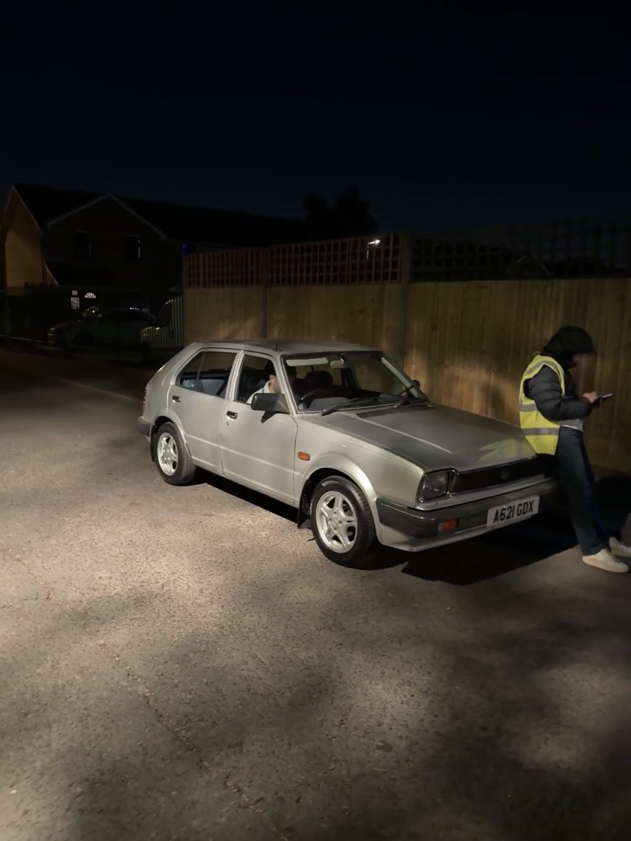 Old silver hatchback parked on a road at night, a man in a hi-vis vest leaning against the bonnet looking at his phone.