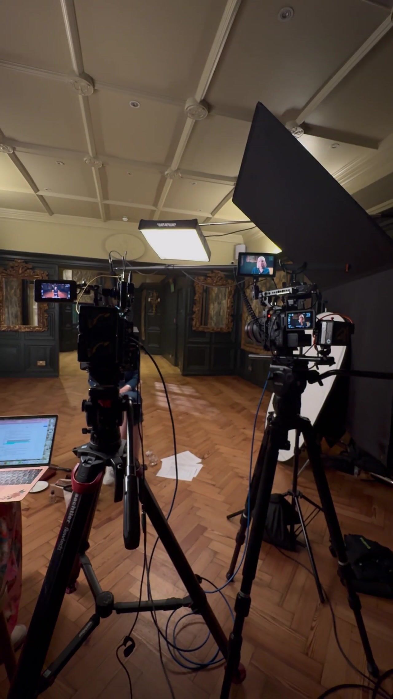 Two cameras on tripods with monitors showing a woman on-screen, facing a lit set with dark panelled walls and gilt-framed mirrors, laptop in foreground.