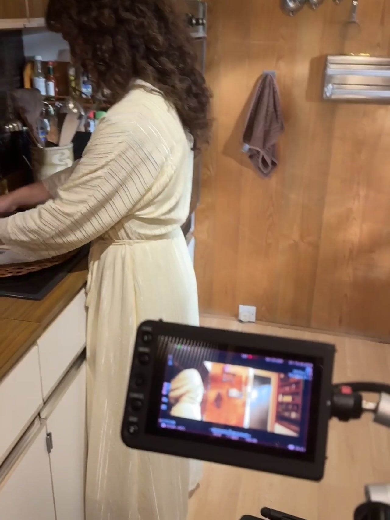 A curly-haired woman in a cream robe works at a kitchen counter, seen past a camera monitor showing the same shot.