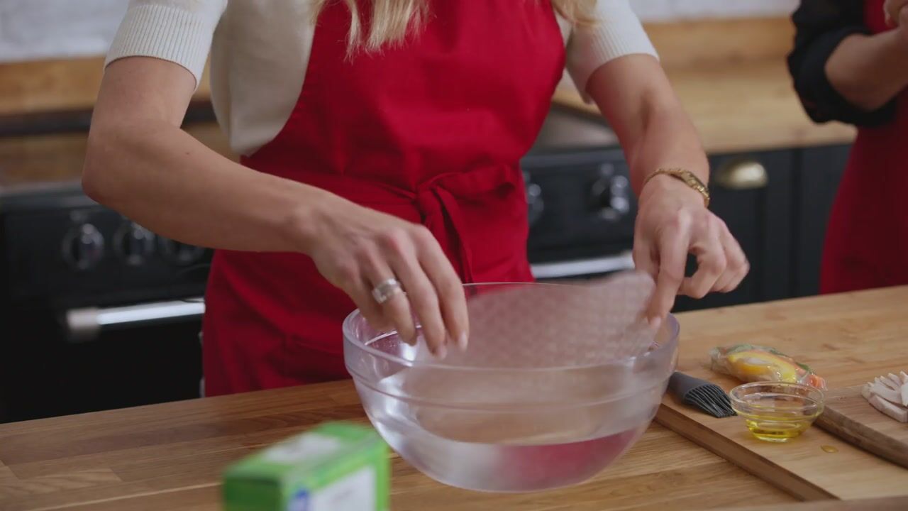 Close-up of a blonde woman in a red apron mixing water in a glass bowl on a wooden worktop, with a lemon and small bowl of oil nearby.