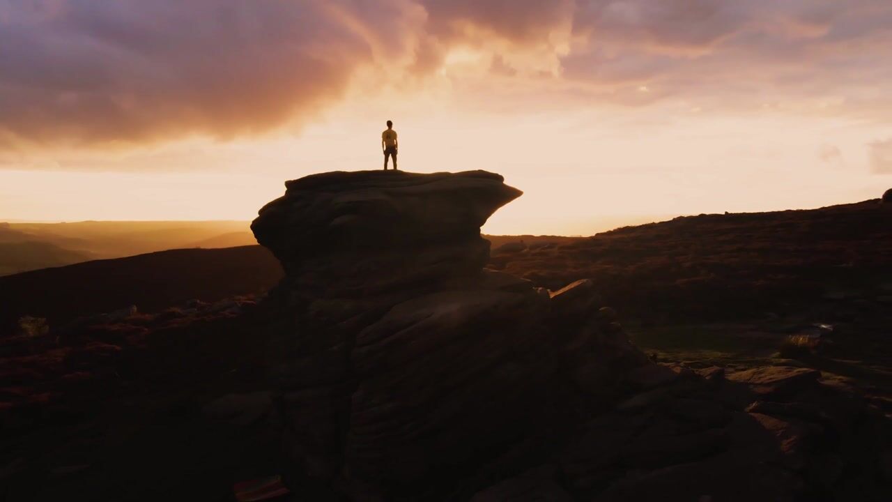 Silhouetted figure standing atop a rocky outcrop on moorland, wide shot against a glowing orange and grey sunset sky.