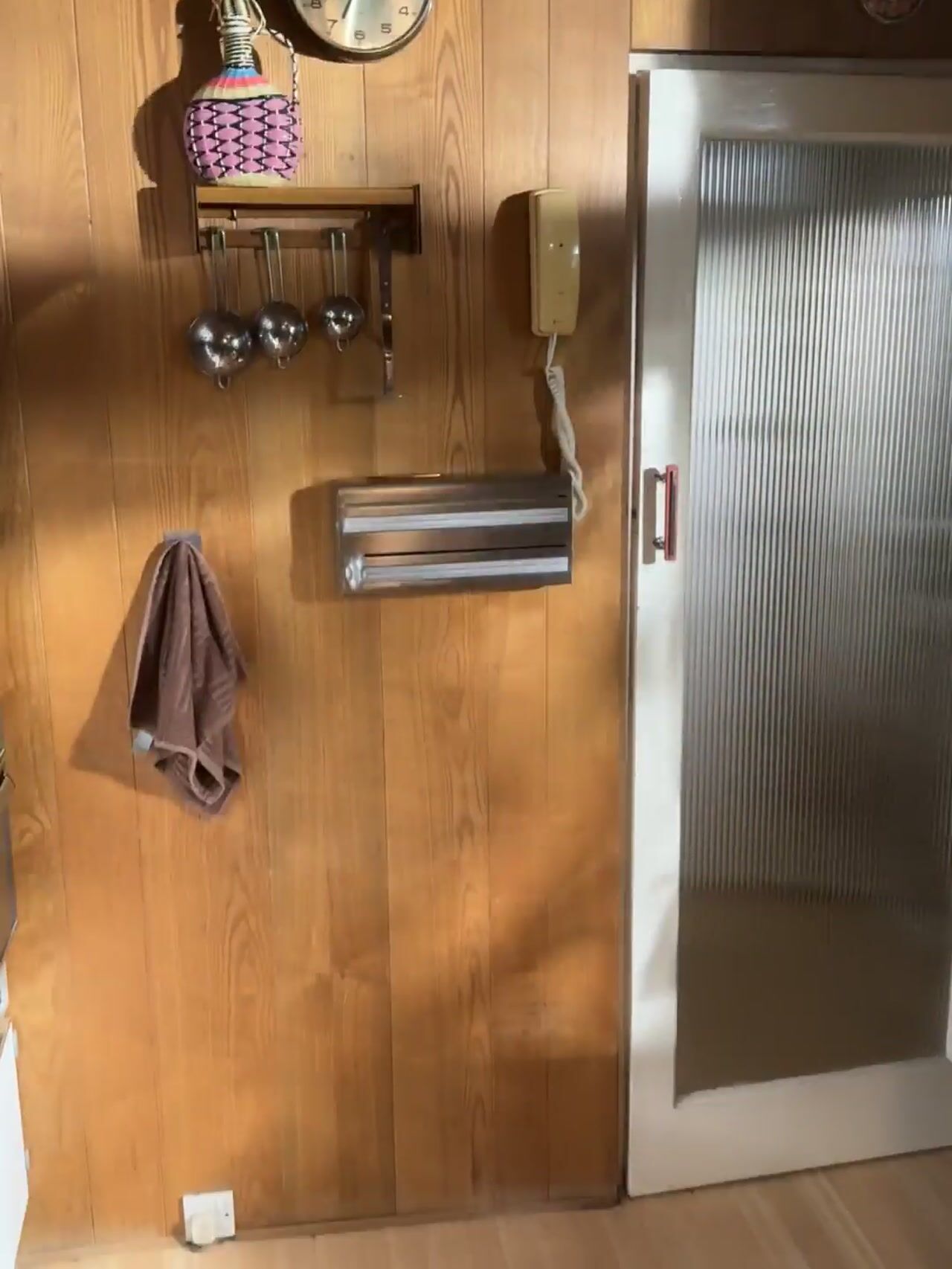 A wood-panelled hallway with a corded telephone, hanging measuring cups, a towel and a paper towel dispenser, beside a frosted-glass door.