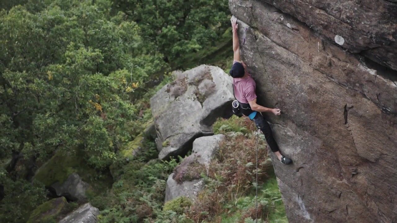 A climber in a pink t-shirt and dark cap scales a rock face, roped, with wooded hillside and boulders visible below.