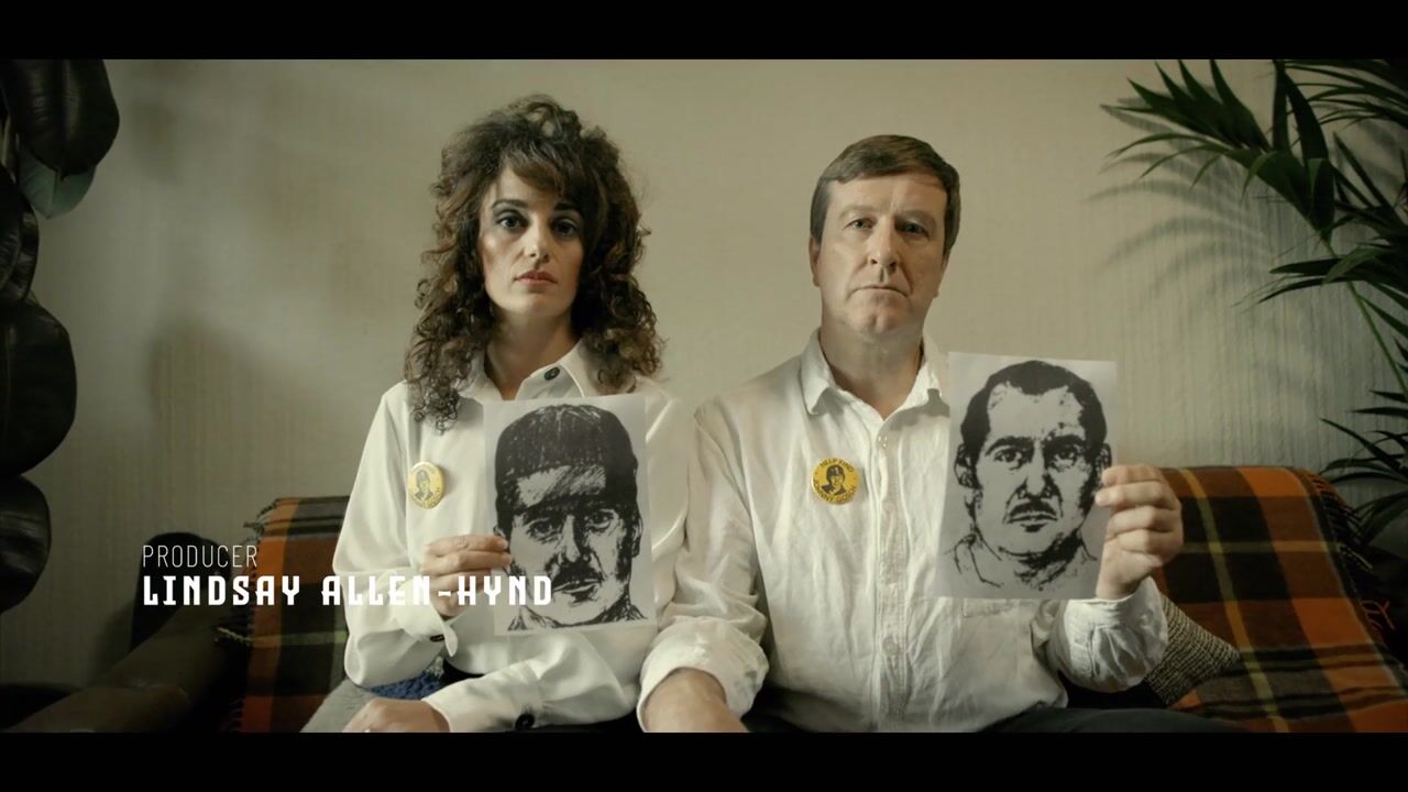 A curly-haired woman and a grey-haired man sit side by side on a plaid sofa, each holding up a sketched portrait, facing camera.