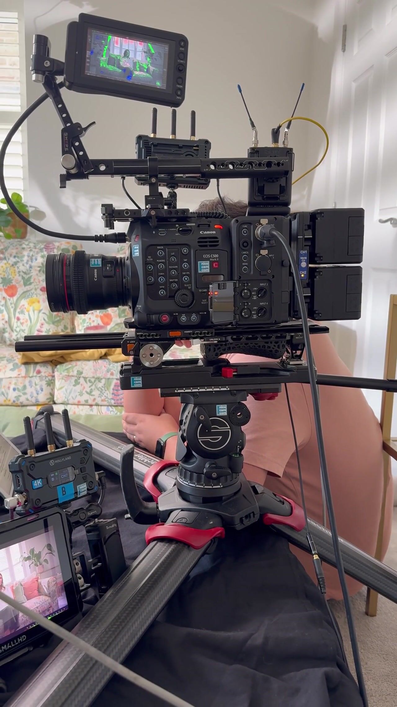 A rigged Canon cinema camera on a tripod with monitors, wireless receivers and cables, framing a floral sofa in a domestic room with shuttered windows.