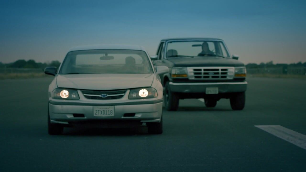 A pale saloon and a dark pickup parked front-to-front on a road at dusk, headlights on, with fields and trees behind.