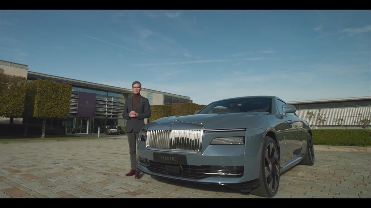 A short-haired man in a grey suit stands beside a grey saloon car labelled "Spectre", facing camera outside a modern building.