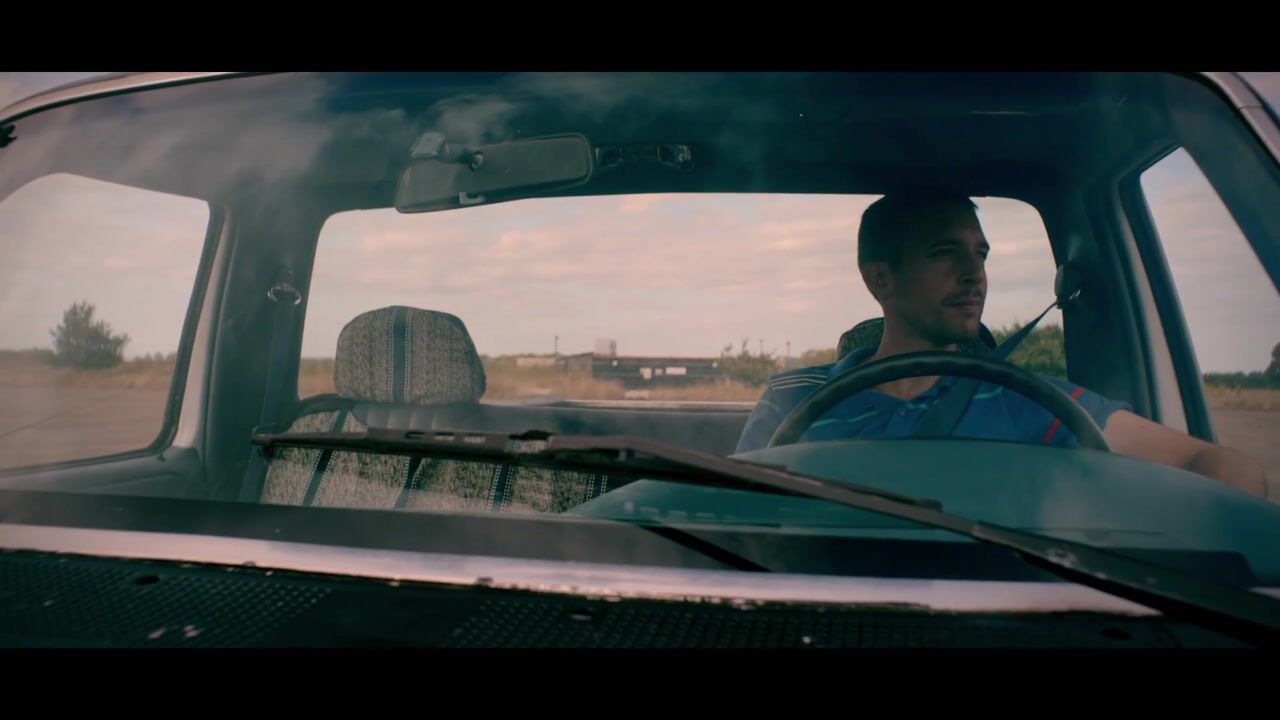 A short-haired man in a blue top drives an old car, seen through the windscreen, with open fields visible outside.