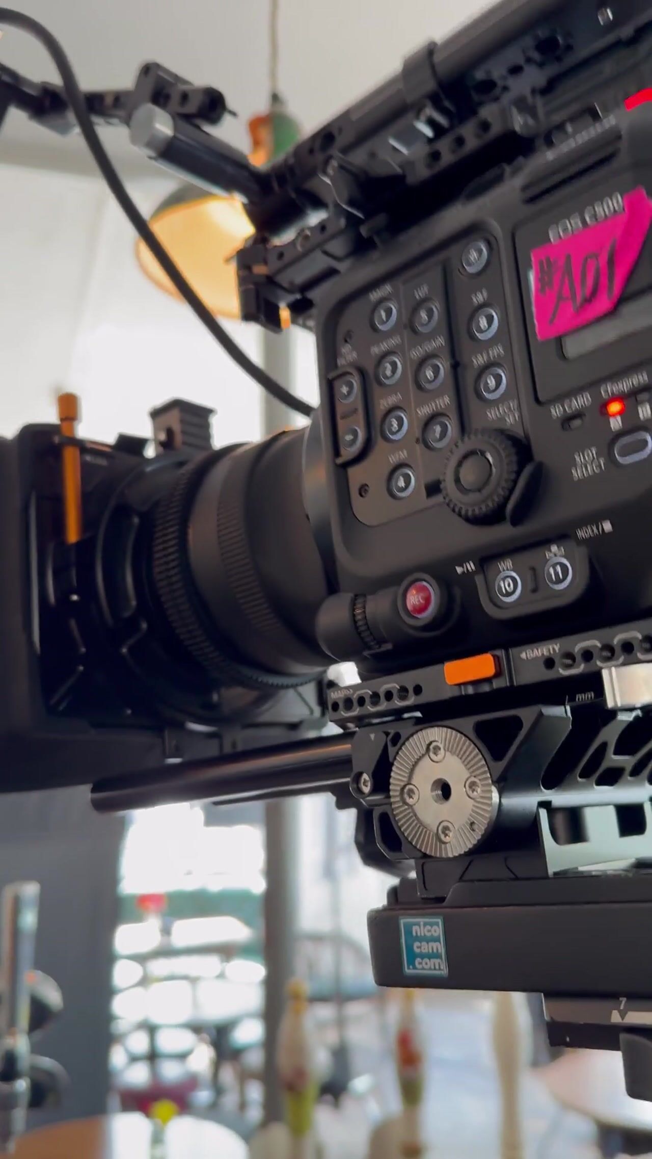 Close-up of a cinema camera's control panel and lens rigged on rails, with a blurred interior and window light behind.